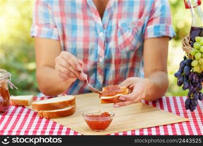 Women&rsquo;s hands spread jam on piece of bread. Red tablecloth, picnic basket, grapes. Natural green background. Sunny day. Picnic concept. Women&rsquo;s hands spread jam on piece of bread