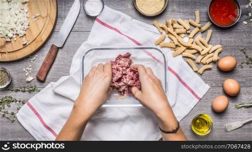 women&rsquo;s hands stuffing mix in a glass bowl, penne pasta, eggs, biscuits panirovachnye, seasonings on wooden rustic background top view close up