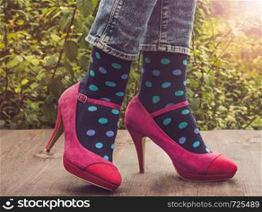 Women's legs in fashionable shoes, bright, multi-colored socks on a wooden terrace on the background of green trees and sunny rays. Close-up. Concept of Style, Fashion and Beauty. Women's legs, bright socks. Concept of Style