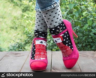 Women's legs in stylish shoes, bright, variegated, multi-colored socks on the wooden terrace on the background of green trees. Lifestyle, fashion, beauty, fun. Women's legs in stylish shoes, bright, variegated, multi-colored socks