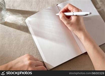 Women’s hand writing in a blank notebook sheet with sunlight shadows, home office lifestyle workspace, mock up with copy space. Women’s hand writing in blank notebook sheet with sunlight shadows, home office lifestyle workspace, mock up with copy space