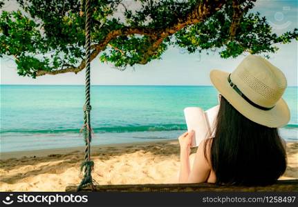 Women sit and reading a book on swings under the tree by the sea. Back view of sexy Asian woman with straw hat relax and enjoy holiday at tropical paradise sand beach. Summer vacation. Summer vibes.