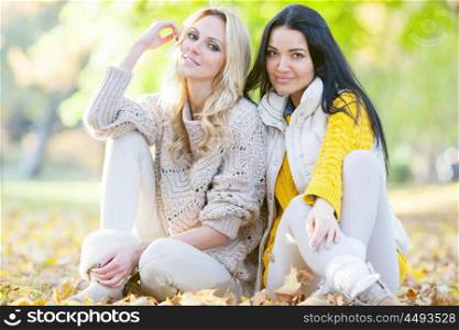 Women sitting in autumn park. Two cheerful women sitting on dry leaves in autumn park at sunny day