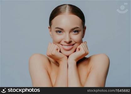 Women, skin care and tenderness concept. Young female takes care of skin and body health, keeps hands under chin and smiles gently, wears makeup, has combed hair, delighted with skin care product