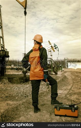 Women worker in the oil field, with wrenches in a hands, orange helmet and work clothes. Industrial site background. Toned.