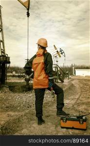 Women worker in the oil field, with wrenches in a hands, orange helmet and work clothes. Industrial site background. Toned.