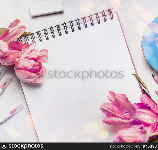 Womens springtime workspace with pale pink tulips, notebook or sketchbook and colorful brush markers , top view