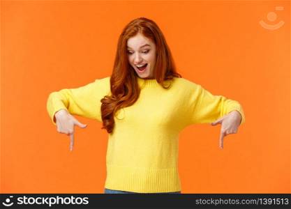 Wondered and excited, amused cute redhead woman in yellow sweater pointing fingers down, look bottom and smiling, check-out product promo, winter holiday event, orange background.. Wondered and excited, amused cute redhead woman in yellow sweater pointing fingers down, look bottom and smiling, check-out product promo, winter holiday event, orange background
