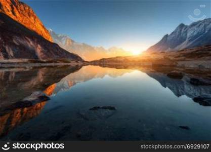 Wonderful landscape with high rocks with illuminated peaks, stones in mountain lake, reflection, blue sky and yellow sunlight in sunrise. Nepal. Amazing scene with Himalayan mountains. Himalayas