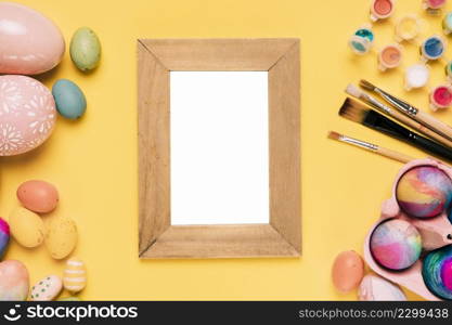 wooden blank white frame with easter eggs paint brushes watercolor paint yellow background