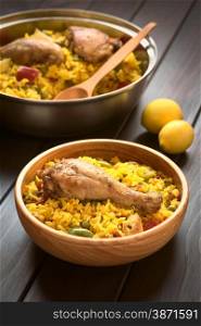 Wooden bowl and a pot of chicken paella, a traditional Valencian (Spanish) rice dish made of rice, chicken, peas and capsicum , photographed on dark wood with natural light (Selective Focus, Focus on the chicken thigh)