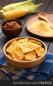 Wooden bowl of homemade baked corn chips with cobs of corn, cornmeal and a bowl of chili con carne in the back, photographed with natural light (Selective Focus, Focus on the front of the chip on the top)