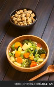 Wooden bowl of vegetable soup made of zucchini, green bean, carrot, broccoli, potato and pumpkin with a small bowl of croutons in the back, photographed on dark wood with natural light (Selective Focus, Focus one third into the soup)