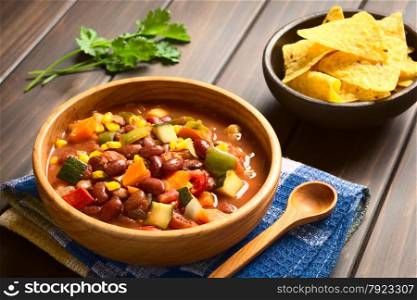 Wooden bowl of vegetarian chili dish made with kidney bean, carrot, zucchini, bell pepper, sweet corn, tomato, onion, garlic, with tortilla chips in the back, photographed with natural light (Selective Focus, Focus in the middle of the dish)