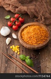 Wooden bowl with boiled red long grain basmati rice with vegetables on wooden table background with sticks and tomatoes with corn,garlic and basil.