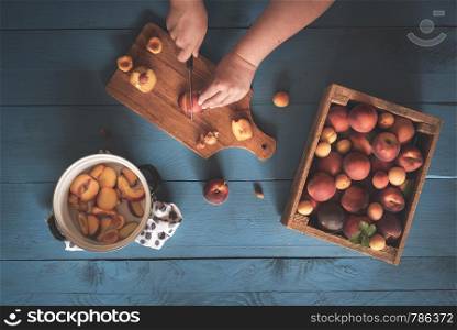 Wooden box with peaches and apricots and a woman hands cutting fruits on a cutting board and in a pot, on a blue table. Flat lay of home cooking.