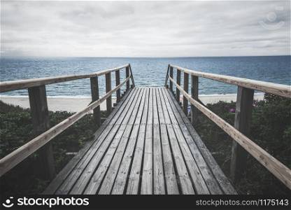 Wooden bridge over roses bushes towards beach and North sea water, on Sylt island, Germany, on a cloudy moody day of summer. Gloomy beach landscape.