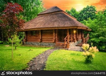 Wooden cottage on the lawn. A house with a path made of sawn logs and a burning lantern stands in a clearing with green grass against the background of a forest. Wooden cottage on the lawn