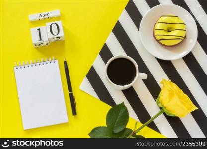 Wooden cubes calendar April 10th. Cup of coffee, yellow donut and rose on black and white napkin, empty open notepad for text on yellow background. Concept stylish workplace Top view Flat lay Mockup. Wooden cubes calendar April 10th. Cup of coffee, yellow donut and rose on black and white napkin, empty open notepad for text on yellow background. Concept stylish workplace