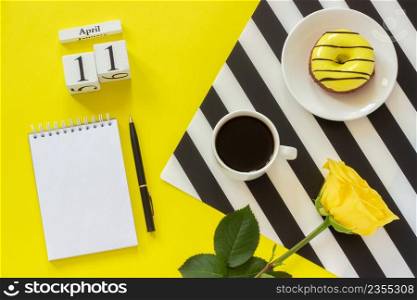 Wooden cubes calendar April 11th. Cup of coffee, yellow donut and rose on black and white napkin, empty open notepad for text on yellow background. Concept stylish workplace Top view Flat lay Mockup. Wooden cubes calendar April 11th. Cup of coffee, yellow donut and rose on black and white napkin, empty open notepad for text on yellow background. Concept stylish workplace