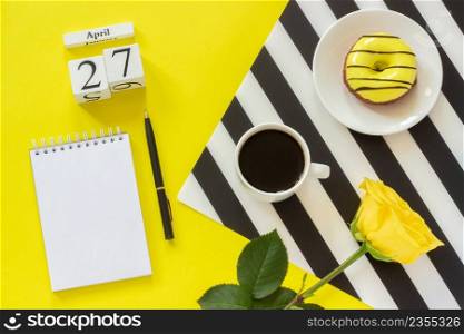 Wooden cubes calendar April 27th. Cup of coffee, yellow donut and rose on black and white napkin, empty open notepad for text on yellow background. Concept stylish workplace Top view Flat lay Mockup. Wooden cubes calendar April 27th. Cup of coffee, yellow donut and rose on black and white napkin, empty open notepad for text on yellow background. Concept stylish workplace