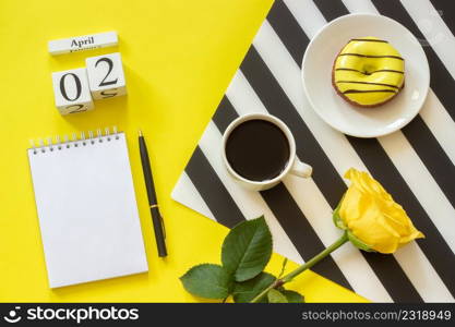 Wooden cubes calendar April 2nd. Cup of coffee, yellow donut and rose on black and white napkin, empty open notepad for text on yellow background. Concept stylish workplace Top view Flat lay Mockup. Wooden cubes calendar April 2nd. Cup of coffee, yellow donut and rose on black and white napkin, empty open notepad for text on yellow background. Concept stylish workplace