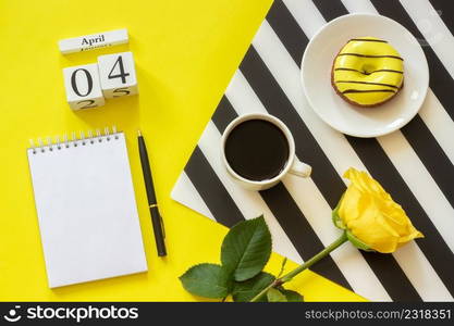 Wooden cubes calendar April 4th. Cup of coffee, yellow donut and rose on black and white napkin, empty open notepad for text on yellow background. Concept stylish workplace Top view Flat lay Mockup. Wooden cubes calendar April 4th. Cup of coffee, yellow donut and rose on black and white napkin, empty open notepad for text on yellow background. Concept stylish workplace