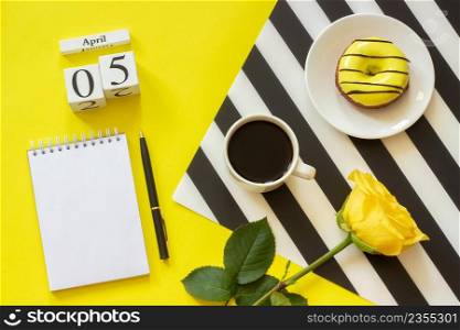 Wooden cubes calendar April 5th. Cup of coffee, yellow donut and rose on black and white napkin, empty open notepad for text on yellow background. Concept stylish workplace Top view Flat lay Mockup. Wooden cubes calendar April 5th. Cup of coffee, yellow donut and rose on black and white napkin, empty open notepad for text on yellow background. Concept stylish workplace