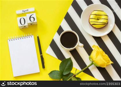 Wooden cubes calendar April 6th. Cup of coffee, yellow donut and rose on black and white napkin, empty open notepad for text on yellow background. Concept stylish workplace Top view Flat lay Mockup. Wooden cubes calendar April 6th. Cup of coffee, yellow donut and rose on black and white napkin, empty open notepad for text on yellow background. Concept stylish workplace
