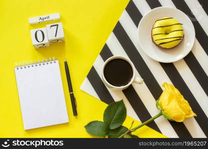 Wooden cubes calendar April 7th. Cup of coffee, yellow donut and rose on black and white napkin, empty open notepad for text on yellow background. Concept stylish workplace Top view Flat lay Mockup. Wooden cubes calendar April 7th. Cup of coffee, yellow donut and rose on black and white napkin, empty open notepad for text on yellow background. Concept stylish workplace