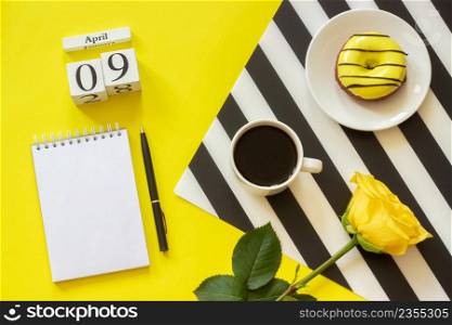 Wooden cubes calendar April 9th. Cup of coffee, yellow donut and rose on black and white napkin, empty open notepad for text on yellow background. Concept stylish workplace Top view Flat lay Mockup. Wooden cubes calendar April 9th. Cup of coffee, yellow donut and rose on black and white napkin, empty open notepad for text on yellow background. Concept stylish workplace