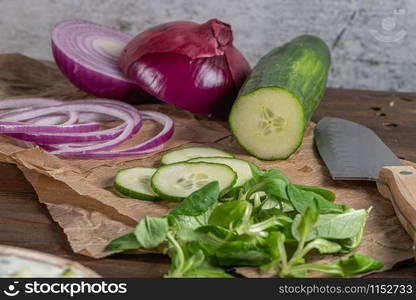 Wooden cutting board on old wooden table top with tablecloth, sliced red onion and cucumber and corn salad