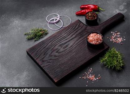 Wooden cutting board, salt, pepper, spices and herbs on a dark concrete background. Cooking delicious home-cooked food. Wooden cutting board, salt, pepper, spices and herbs on a dark concrete background