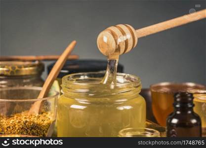 Wooden dipper with honey flowing into glass jar. Various types of honey, honey bee pollen, propolis and wooden honey dipper on table. . Honey Flowing into a Glass Jar