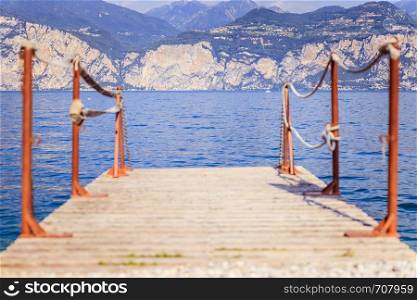 Wooden dock pier extending over blue lake water, mountains at lago di garda. Text space.