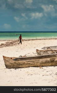 Wooden dugout boats lie on the beach sand of Zanzibar near the clear blue and aqua waters of the Indian ocean with some heavy clouds approaching In the distance an unrecognizable person crosses the white beach sand.