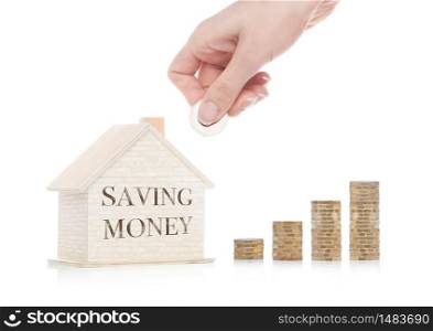 Wooden house model with coins next to it and hand holding the coin with conceptual text. Saving Money