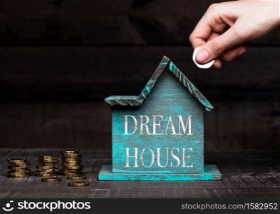 Wooden house model with coins next to it and hand holding the coin with conceptual text. Dream House