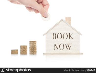 Wooden house model with coins next to it and hand holding the coin with conceptual text.Book Now