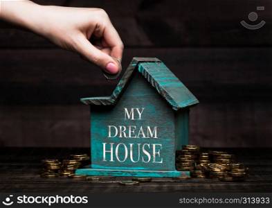 Wooden house model with coins next to it and hand holds the coin with conceptual text. My Dream House