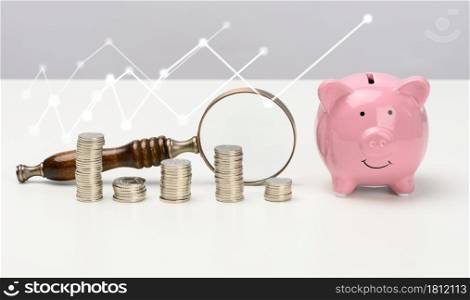 wooden magnifier and pink ceramic piggy bank and a stack of coins on a white table. Savings concept, budget planning. Income growth