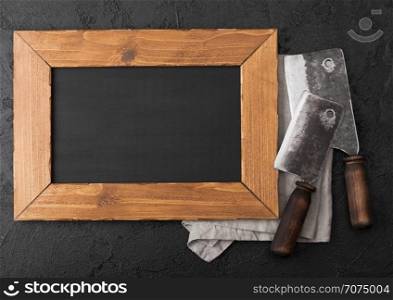 Wooden menu board and vintage meat knife hatchets on kitchen towel and black stone table background. Butcher utensils.