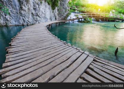 Wooden path across beautiful lake and waterfall in sunny green forest
