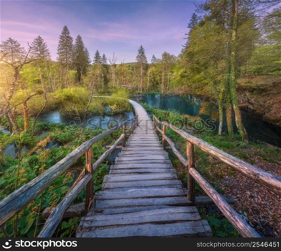 Wooden path in green forest in Plitvice Lakes, Croatia at sunset in spring. Colorful landscape with stairs in blooming park, trees, water lilies, river, pink sky in summer. Trail in woods. Nature