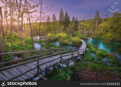 Wooden path in green forest in Plitvice Lakes, Croatia at sunset in spring. Colorful landscape with stairs in blooming park, trees, water lilies, river, pink sky in summer. Trail in woods. Nature