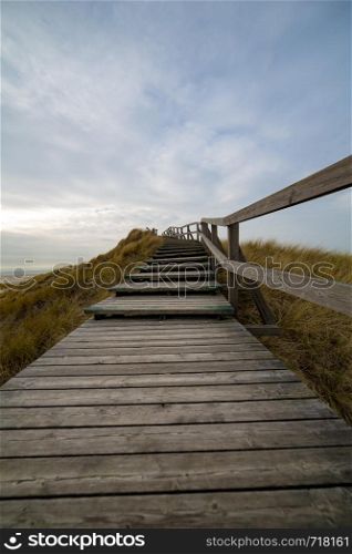 Wooden path with stairs or boardwalk leading through dunes landscape on Amrum to the top of a hill and a new life, North Frisian Islands, Schleswig-Holstein, Germany on a cloudy day