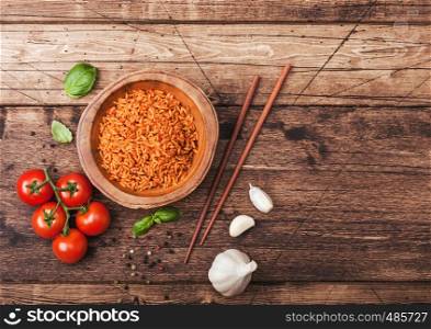 Wooden plate bowl of rice with tomato and basil and garlic and chopsticks on brown bamboo place mat on wood background with dark cloth. Top view.