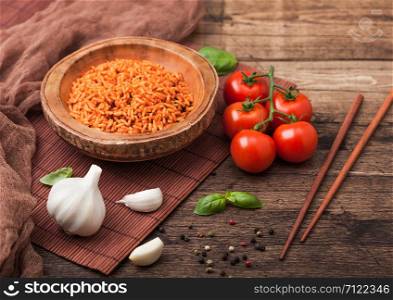 Wooden plate bowl of rice with tomato and basil and garlic and chopsticks on brown bamboo place mat on wood background with dark cloth. Top view.