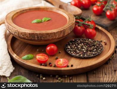 Wooden plate of creamy tomato soup 0n round tray, pepper and kitchen cloth on wooden board with box of raw tomatoes.