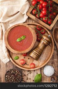 Wooden plate of creamy tomato soup 0n round tray, pepper and kitchen cloth on wooden board with box of raw tomatoes. Top view